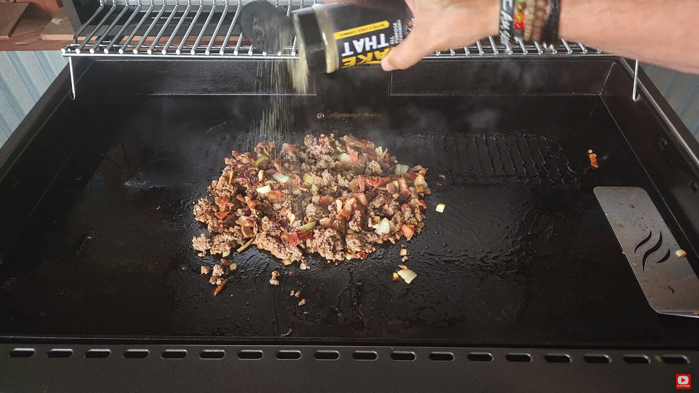 Ground beef, bacon, and  onions on a griddle with seasoning being sprinkled over them